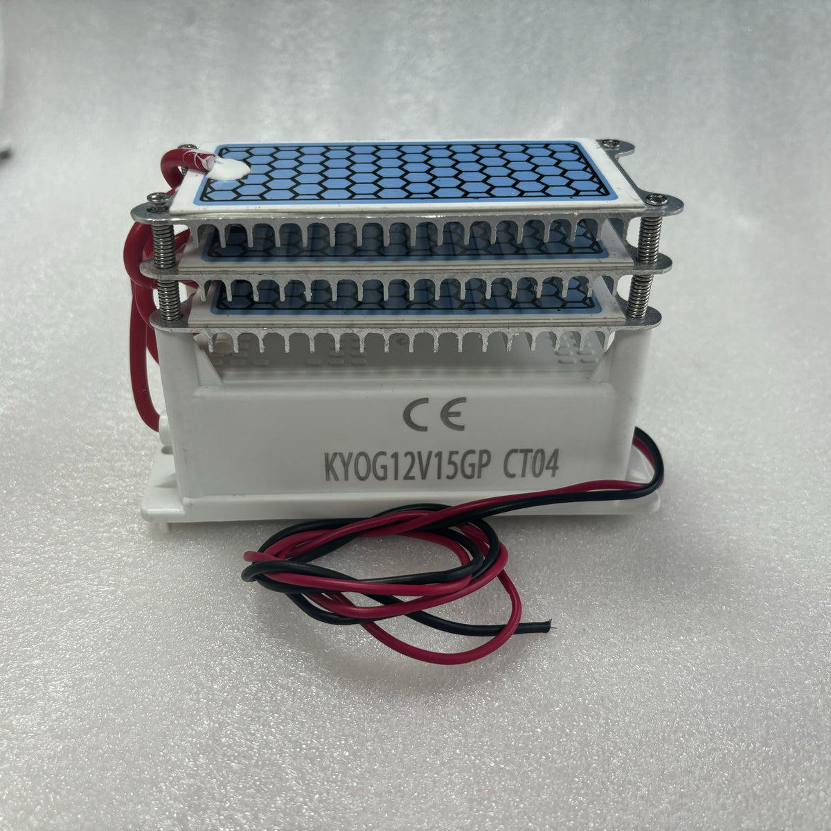    Side view photograph of the KYOG220V15GP CT04 ceramic ozone generator module on a clean white surface, showcasing the white flame-retardant housing with official CE marking and model silkscreen, red and black DC power input wires, aluminum cooling bracket, and dual honeycomb dielectric plates.
   